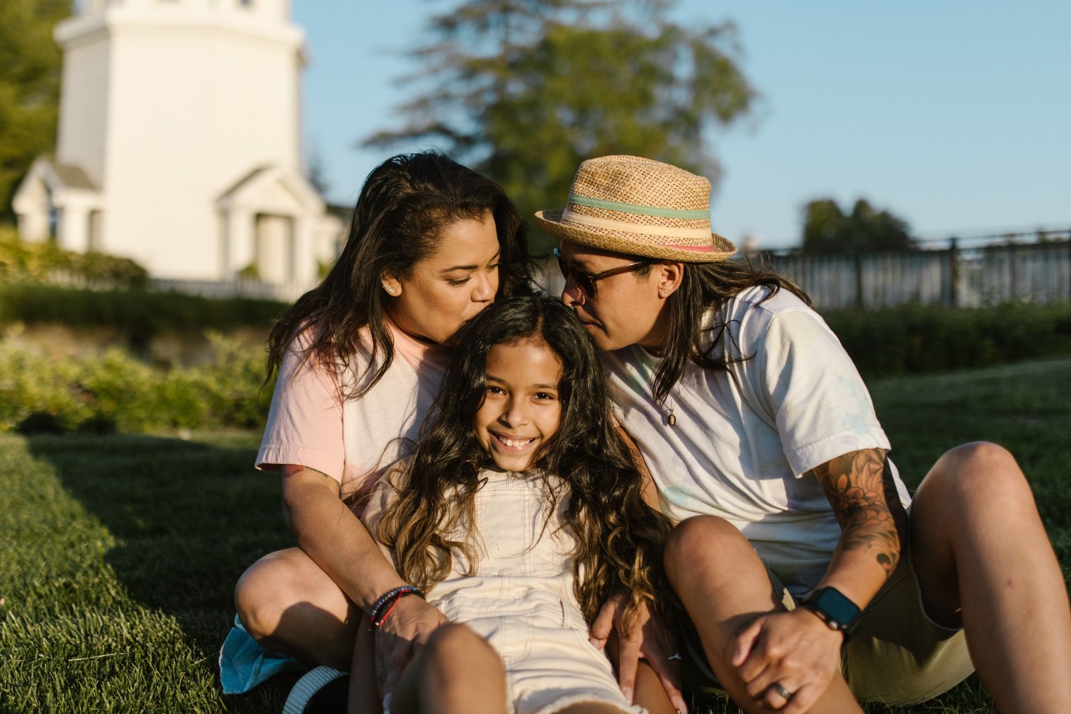 Two adults sit on the grass with a child between them in warm evening light. One kisses the top of the child's head while the child smiles toward the camera.